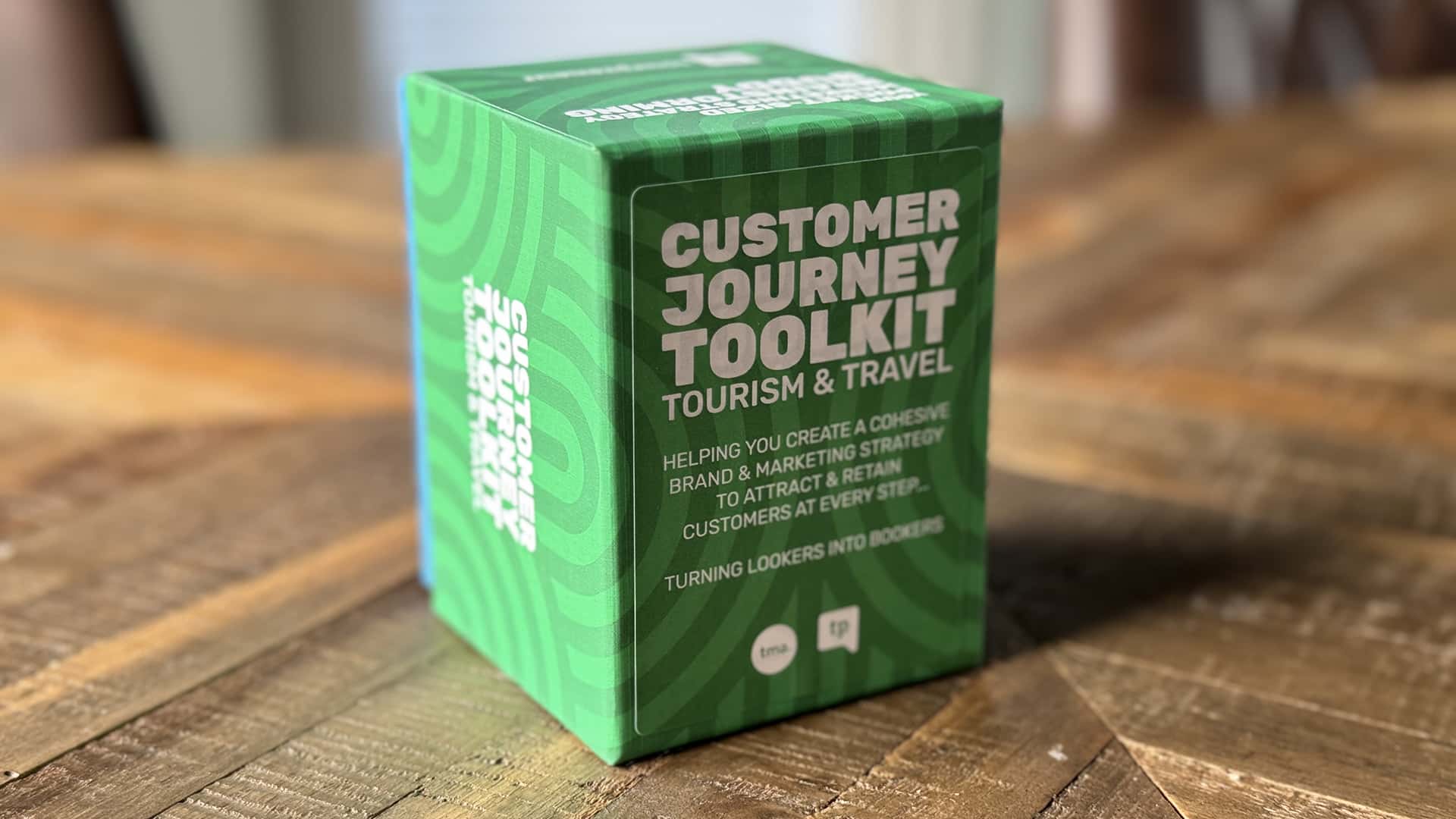 A green box labeled "Customer Journey Toolkit: Tourism & Travel," sits on a wooden surface. The box is designed to help with brand and marketing strategy to attract and retain customers.