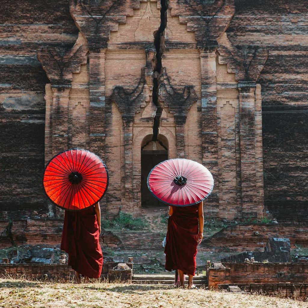 Destination marketing agency by Tourism Marketing Agency, two monks holding red and white umbrellas standing before an ancient temple wall, highlighting cultural travel experiences