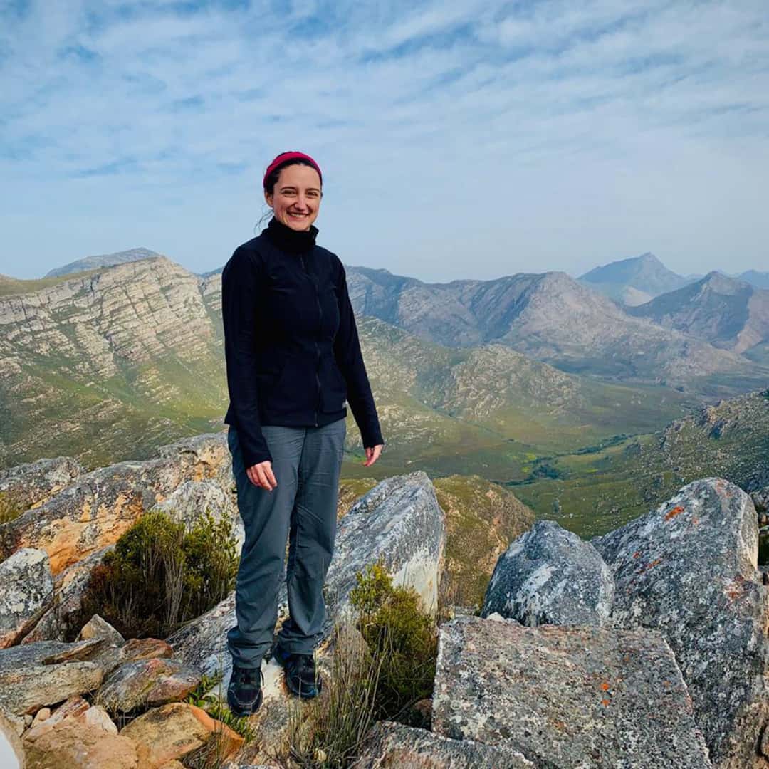 Amy Pieterse, in her sturdy outdoor attire, stands on rocky terrain with a backdrop of majestic mountains and a cloudy sky.