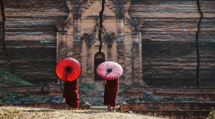 Two individuals in traditional attire hold red umbrellas while standing in front of an ancient, cracked structure made of brick, embodying the kind of dedication and passion we seek. Junior Account Manager required to join our team.