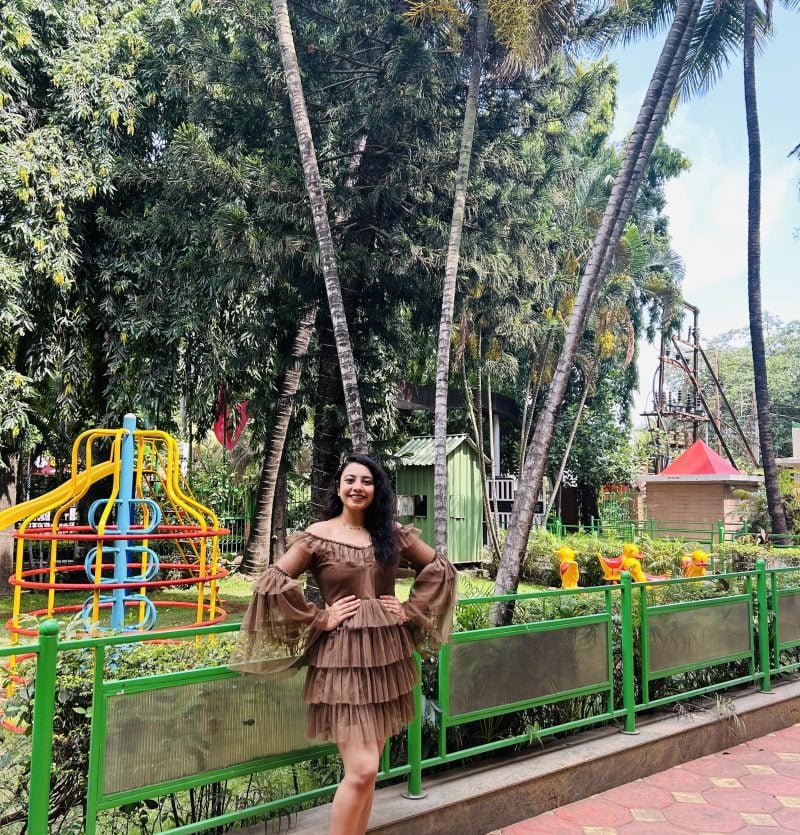 Woman in a brown dress stands smiling on a paved walkway with green railings, trees, and colorful playground equipment in the background, showcasing the confidence of a creative web developer enjoying some time outdoors. Woman in a brown dress stands smiling on a paved walkway with green railings, trees, and colorful playground equipment in the background, showcasing the confidence of a creative web developer enjoying some time outdoors.
