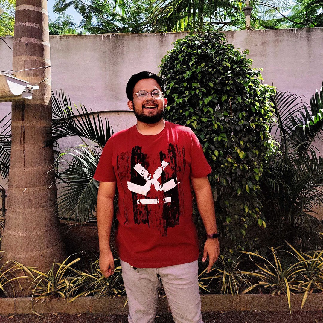 A man with glasses and a beard stands outdoors in front of plants and a wall, wearing a red graphic t-shirt and light pants, smiling at the camera.