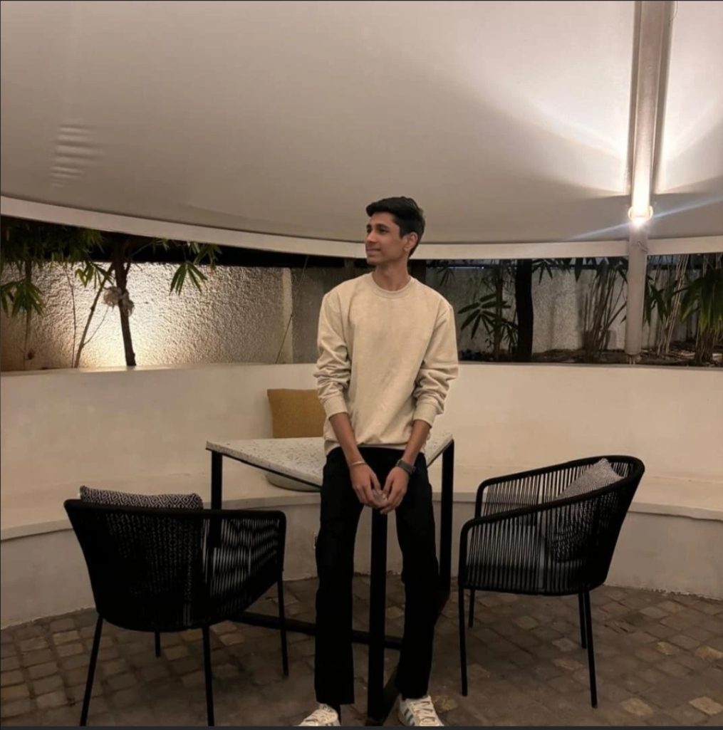 A Social Media Executive stands between two black chairs at an outdoor table under a white canopy, with plants and wall lights in the background.