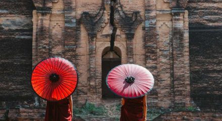 Destination marketing agency by Tourism Marketing Agency, two monks holding red and white umbrellas standing before an ancient temple wall, highlighting cultural travel experiences